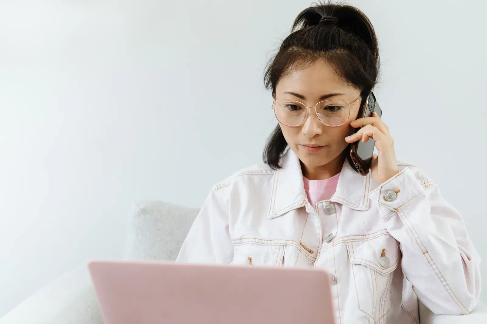 Woman in glasses multitasking with phone and laptop in a serene office setting.